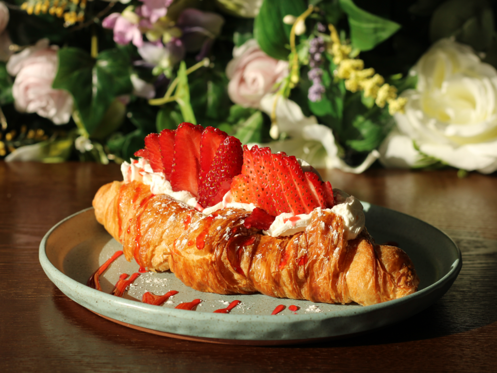 A flaky croissant filled with fresh strawberries, fluffy whipped cream, and sweet strawberry jam, dusted with powdered sugar for a dreamy finish.