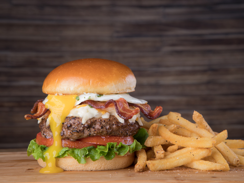 A juicy 1/3 lb burger with an over-easy egg, crispy bacon, fresh lettuce, tomato, bread & butter chips, melted American cheese, and house burger sauce, all served on a toasted brioche bun.